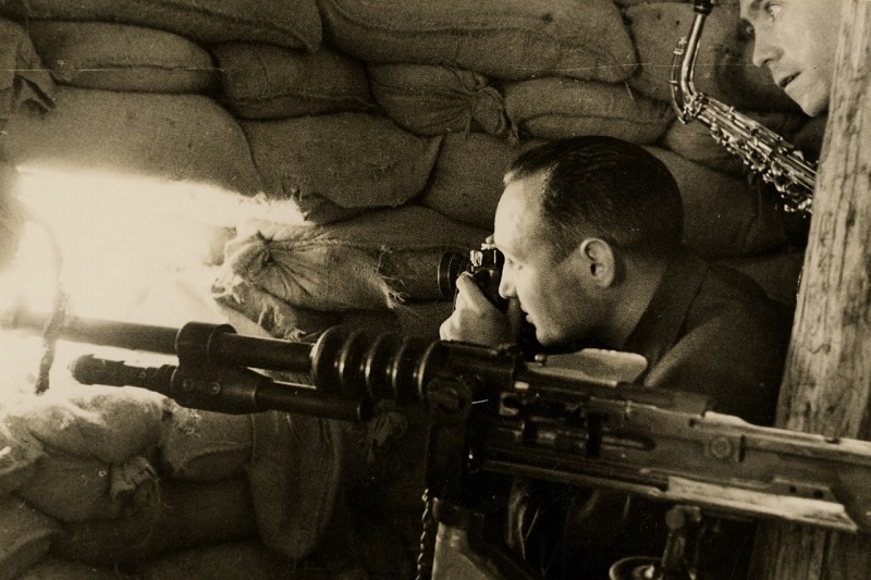 Due miliziani repubblicani, uno con in mano un saxofono e l’altro mentre scatta delle foto attraverso la feritoia, dentro a una postazione fortificata dei ribelli all’interno della caserma militare di Cuartel de la Montañada cui i fascisti sparavano con mitragliatrici di grosso calibro. Madrid (Spagna), 1936